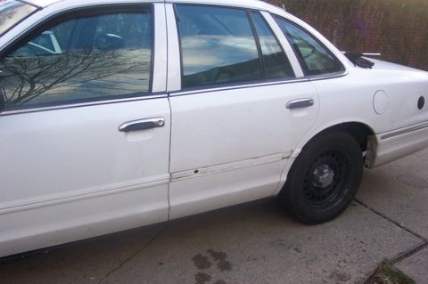 1996 Ford Crown Victoria - The side of a white sedan shows damage along the lower body trim and a black steel wheel.