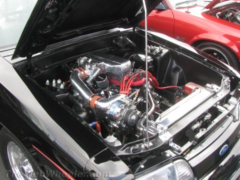 1968 Ford Mustang - The engine bay of a black car is shown, with a prominent supercharger and custom wiring, while a red car is visible in the background.