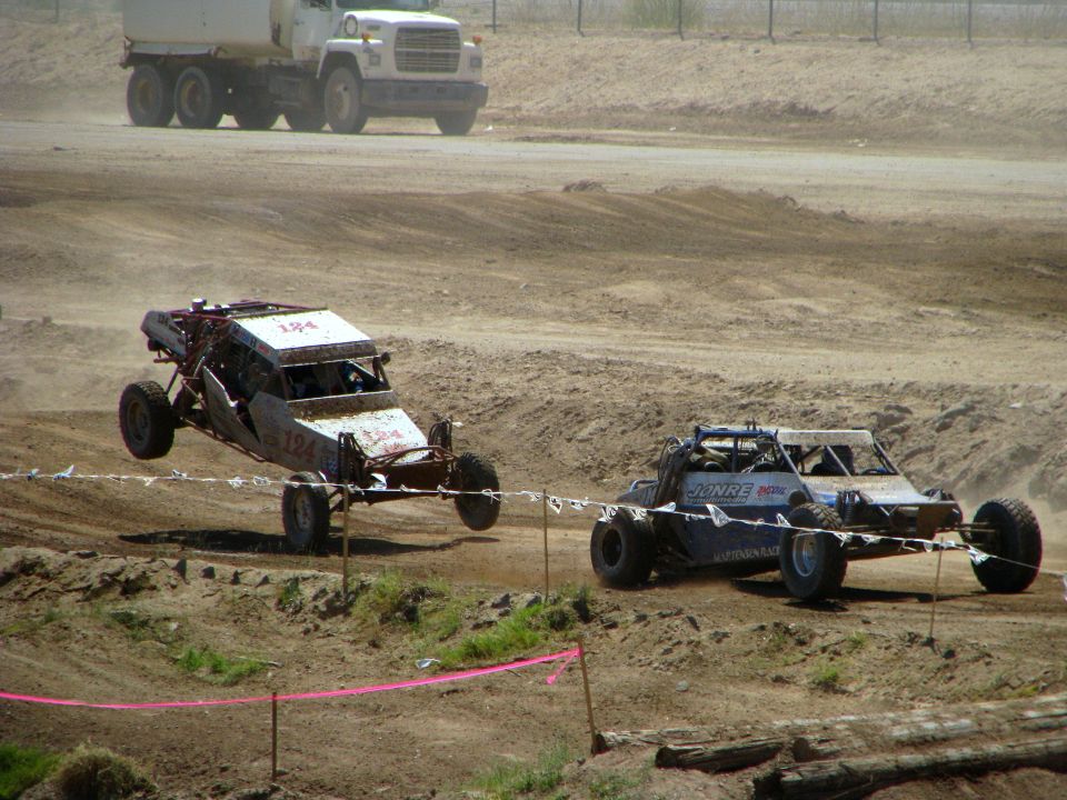 Two off-road buggies race on a dirt track; one is airborne. A large truck is in the background
