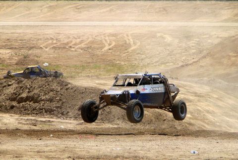 2008 Ford Bronco - An off-road buggy jumps over a dirt mound, with another vehicle partially buried in the dirt in the background.