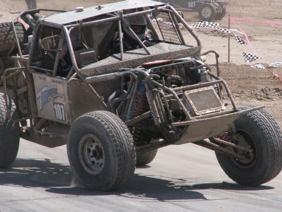 Mud-covered buggy with number 107 on the side, racing on a dirt track. Another buggy is visible in the background