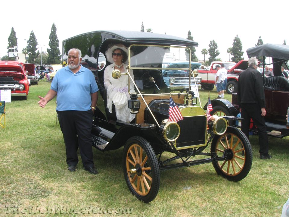 Man stands next to a black antique car with a woman inside. Other cars and people are in the background