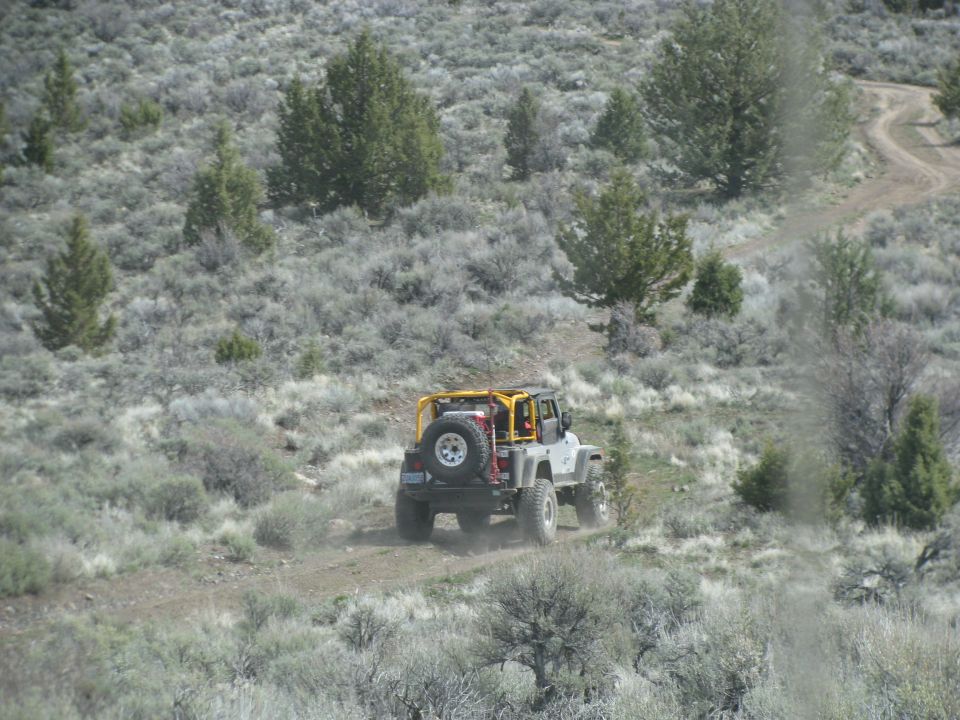 Silver open-top Jeep Wrangler with yellow roll cage and Hi-Lift jack on a desert two-track