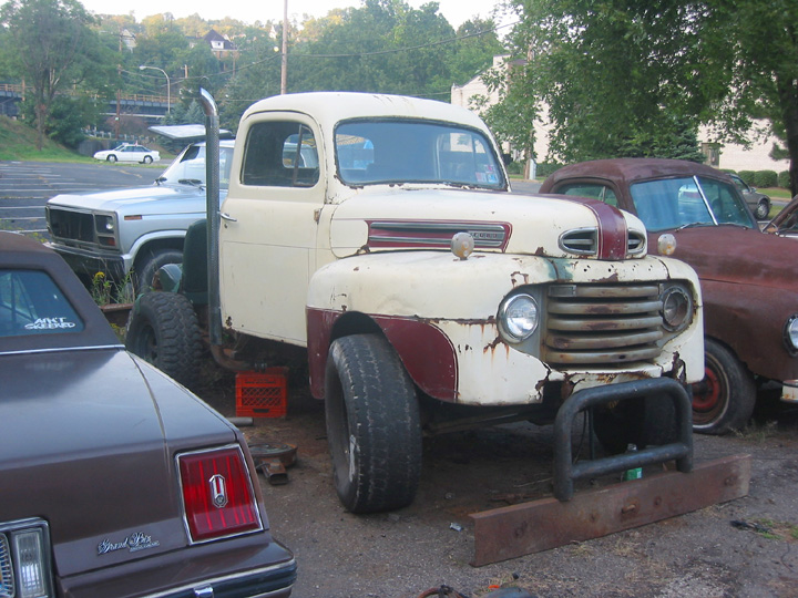 Cream and maroon vintage Ford pickup with rusty grille and tall exhaust stack in a gravel lot