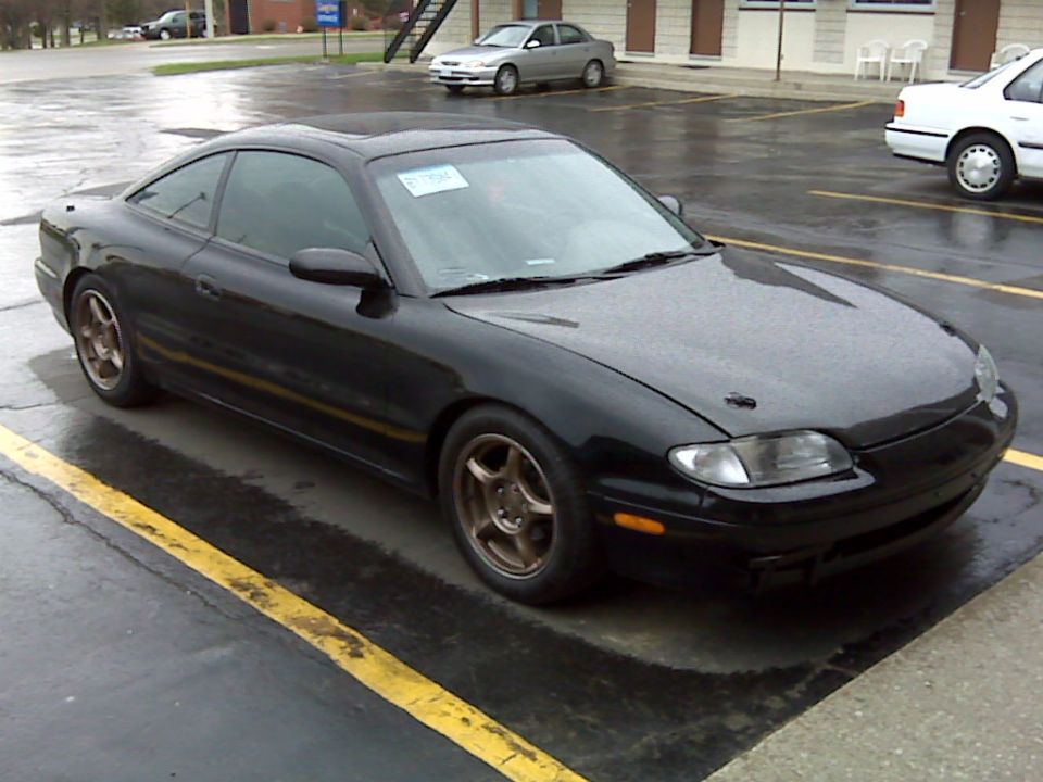 Black 1997 Mazda MX-6 coupe with bronze wheels parked in a wet lot.