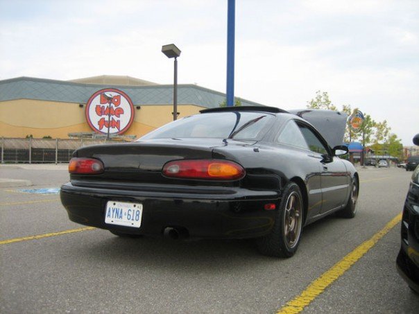 Black 1997 Mazda MX-6 coupe, rear driver's side view, hood open, parked in lot with 'Big Time Fun' sign.