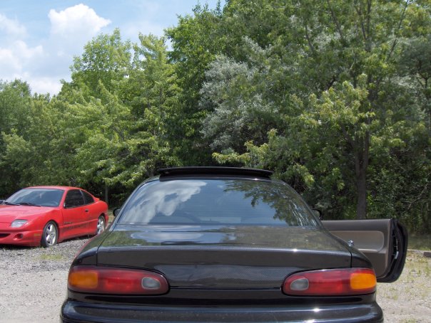 Rear view of a black coupe with driver's door open, red coupe in background.