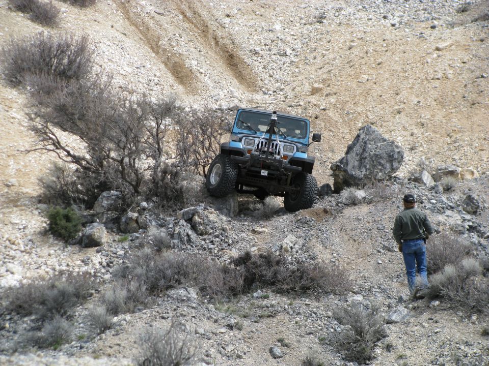 Blue Jeep Wrangler YJ crawling a rocky desert ledge with a man walking below