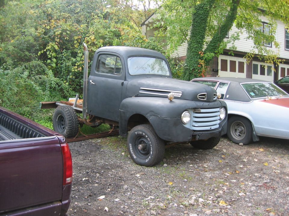 Flat black vintage Ford F1 pickup with bare frame instead of bed, parked on gravel beside other vehicles