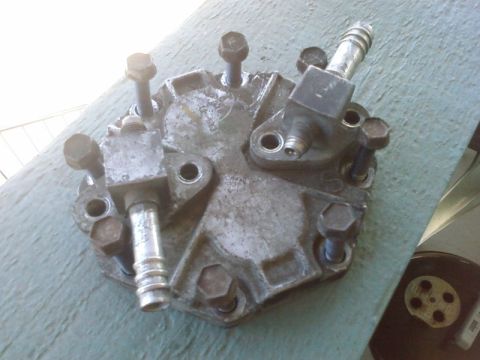 1995 Ford Bronco - A close-up, overhead view of a metallic, possibly aluminum, automotive part with several bolts and two hose barb fittings. The part is weat…