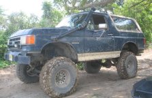 1990 Ford Bronco - A dark blue, lifted 1990 Ford Bronco is parked on a dirt path with large off-road tires. The vehicle has a snorkel intake on the driver's s…