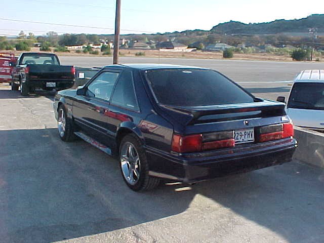 Dark blue Fox-body Mustang hatchback with spoiler and chrome wheels, rear three-quarter view in a parking lot