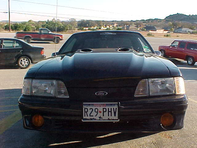 Front view of a black Fox-body Ford Mustang with hood scoops and Texas plate R29-PHV in a parking lot