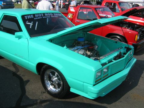 1992 Ford Mustang - A teal coupe is parked with its hood open, revealing the engine bay. A red truck is visible behind it.