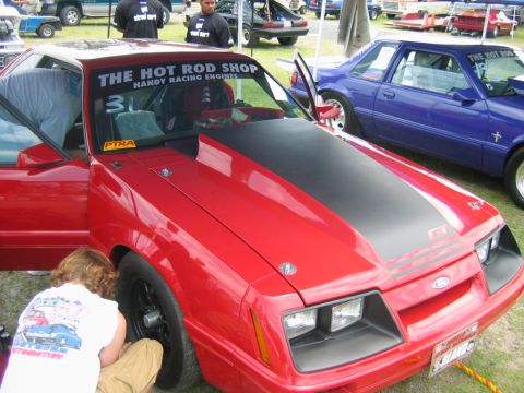 1992 Ford Mustang - A red 1980s Ford Mustang with a prominent black hood stripe and 'GT' badging is parked next to a blue Mustang at an outdoor event.