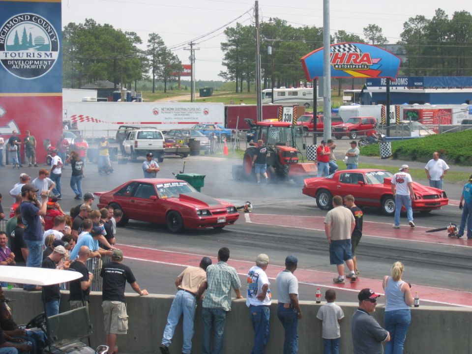 Red Ford Mustang and another red car at a drag strip with spectators and a tractor