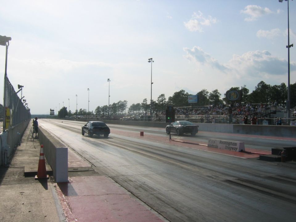 Two cars racing on a drag strip with spectators in the background. Rockingham Dragway sign visible