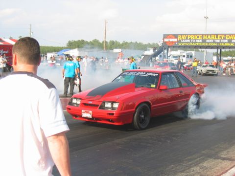 1992 Ford Mustang - A red drag car performs a burnout on the track, surrounded by spectators and other vehicles.