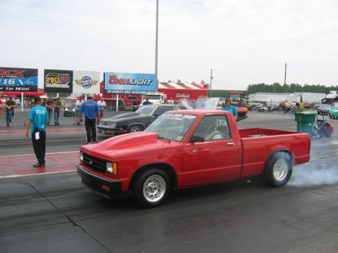 1992 Ford Mustang - A red pickup truck performs a burnout on the drag strip, with spectators and other vehicles in the background.