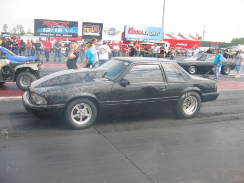 1992 Ford Mustang - A black Ford Mustang sits on the drag strip, ready for a race, with spectators and other vehicles in the background.