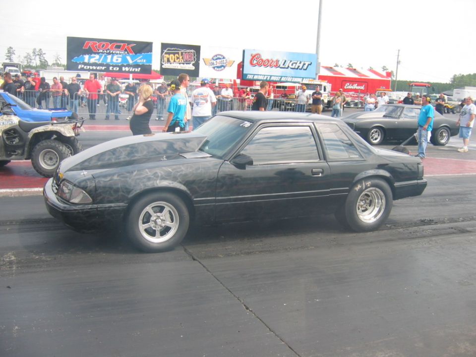 Black drag car with custom hood flames on track, another car and ATV nearby