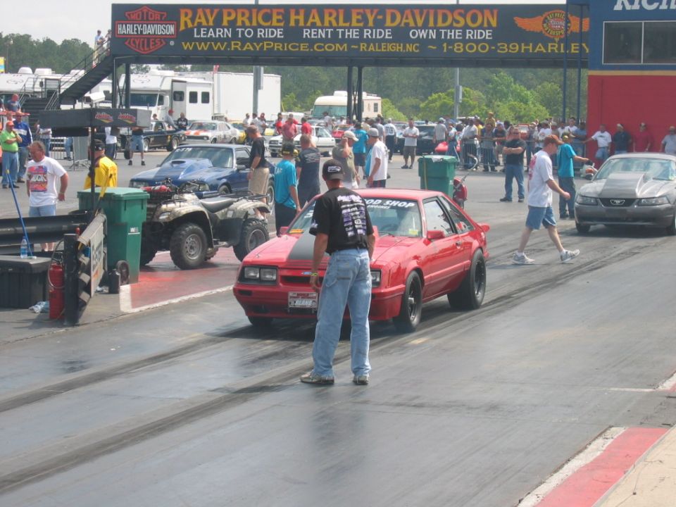 Red Ford Mustang Fox Body with black stripe on hood at drag strip, another car visible on right