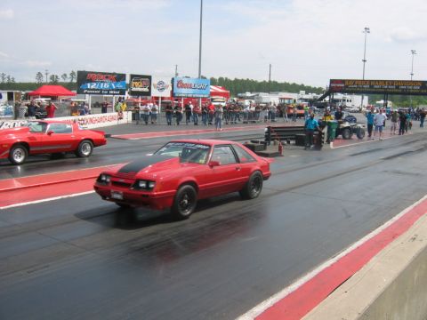 1992 Ford Mustang - A red Ford Mustang races down the drag strip with spectators watching from the sidelines.