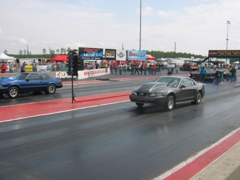1992 Ford Mustang - Two Ford Mustangs race down a drag strip, with spectators and advertising banners in the background.