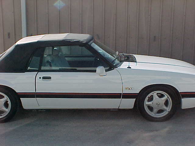 White convertible sports car parked outside, black top up. Side view shows red stripe along the bottom