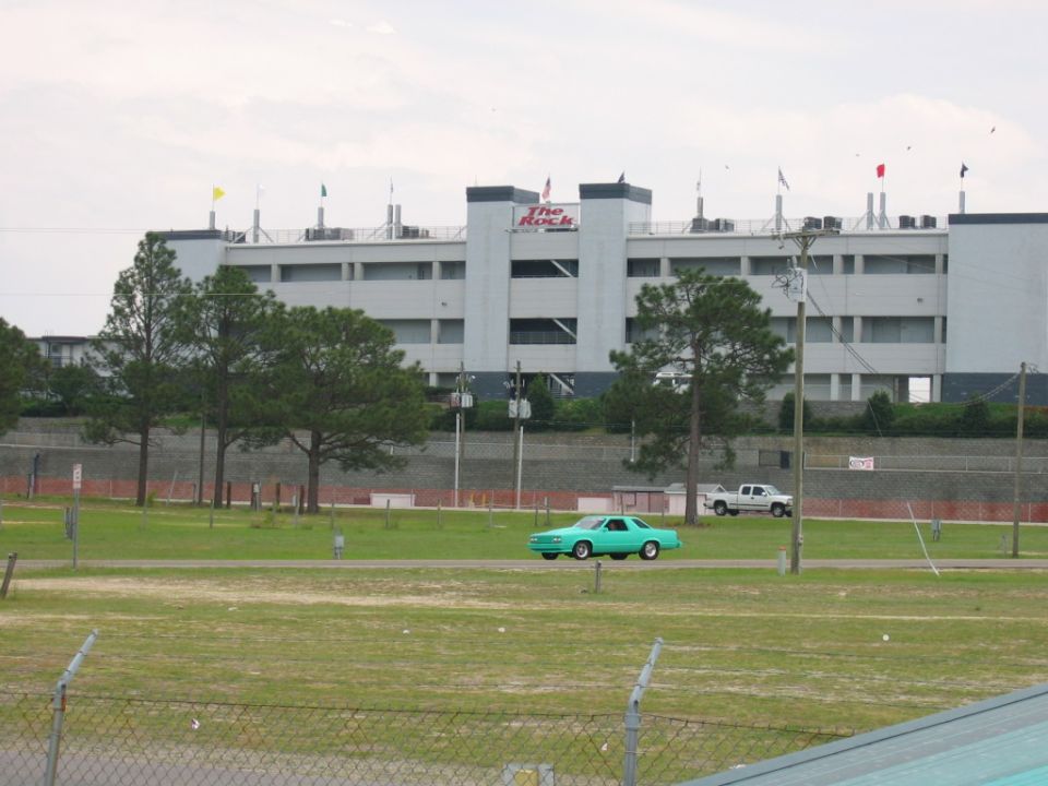 Teal coupe and white pickup truck on a road in front of a grandstand building with 'The Rock' logo
