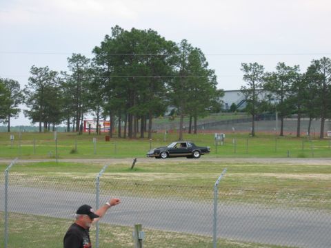 1992 Ford Mustang - A man watches a classic car drive by at a venue with a grandstand and trees.