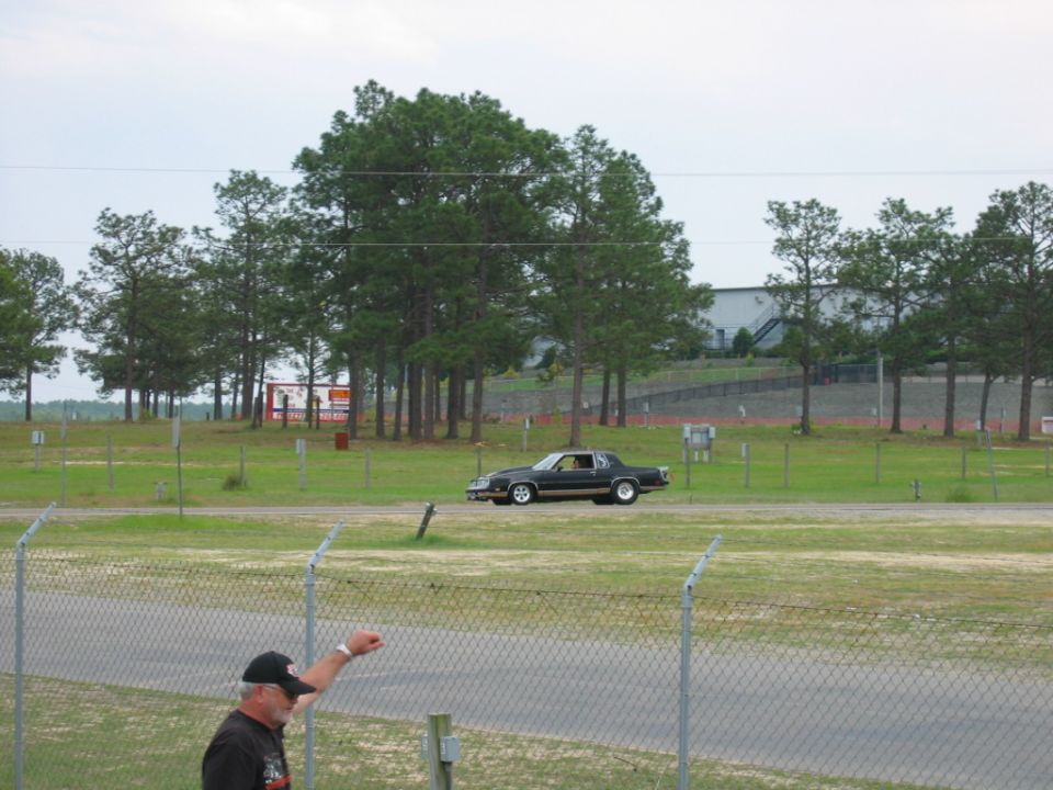 Black coupe drives past trees and a grandstand at a raceway. A man in a cap points towards the track