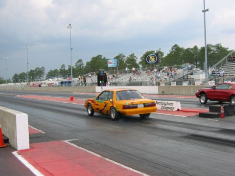 1992 Ford Mustang - An orange fox body Mustang races down a drag strip, with spectators in the stands and a red car in the adjacent lane.