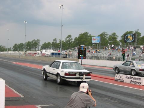 1992 Ford Mustang - Two Mustangs compete on a drag strip under cloudy skies, with a photographer capturing the action.