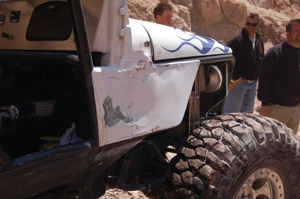 Modified off-road vehicle with white body, blue flame decals, and large mud tires. Three men stand behind it