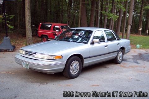 2001 Ford Crown Victoria - A silver sedan is parked in front of a red SUV, with trees in the background. Text indicates it's a 1996 Crown Victoria.