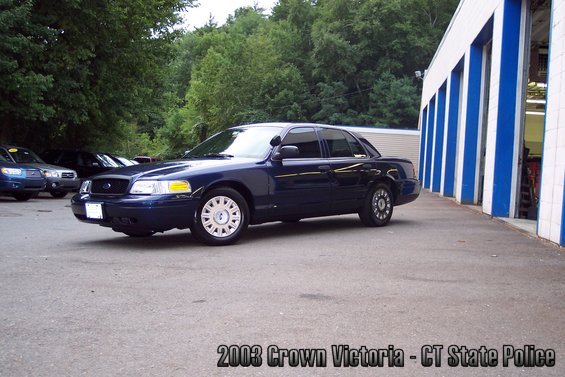 Dark blue 2003 Ford Crown Victoria sedan, front quarter view, parked on asphalt in front of a building with blue and white striped garage doors