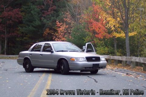 2001 Ford Crown Victoria - A silver 2004 Ford Crown Victoria police car is parked on a road with the driver's door open, surrounded by trees in autumn colors.