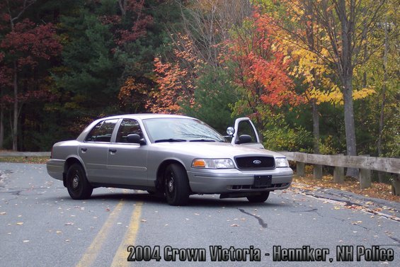 Silver 2004 Ford Crown Victoria police car, driver's door open, parked on a road with autumn foliage