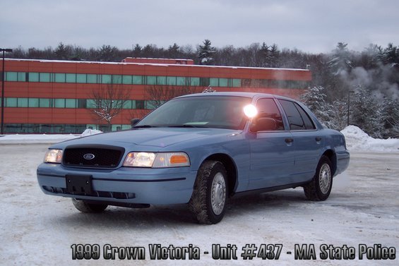 Blue 1999 Ford Crown Victoria police car parked in snow, with a building and trees in the background
