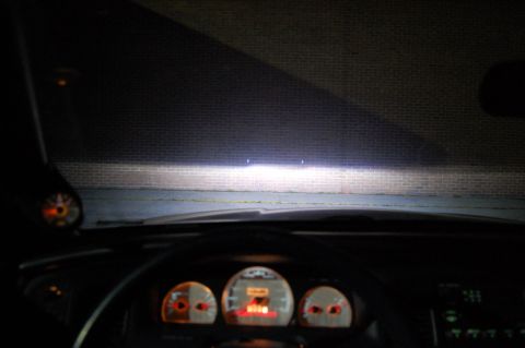 1985 Mercury Grand Marquis - The view from inside a car at night, with illuminated dashboard gauges and headlights shining on a brick wall.