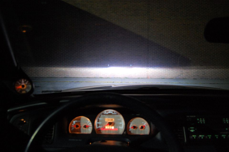 Car interior at night with lit gauge cluster and headlights on a brick wall through the windshield