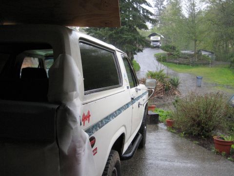 1980 Ford Bronco - A white SUV with a 4x4 decal is parked in a driveway on a rainy day, with an RV visible further up the road.