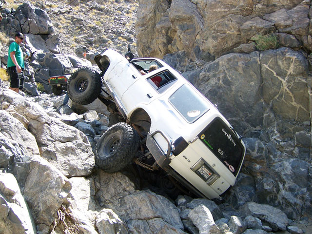 White SUV is tilted on its side, wheels high up on rocks. Another off-road vehicle is behind it. A person stands watching