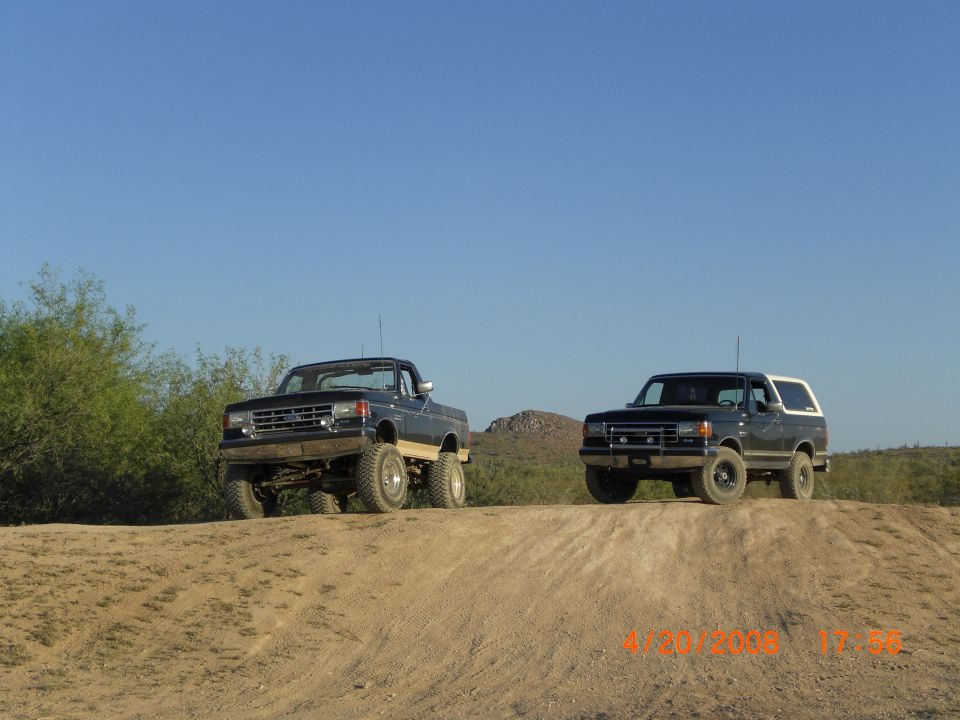 Two dark 1989 Ford Broncos on a dirt hill under a clear blue sky