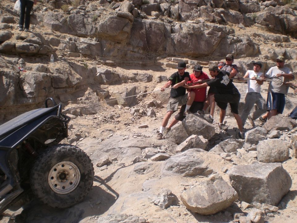 Off-road vehicle's rear tire and roll cage stuck on rocky terrain, people pulling with a rope