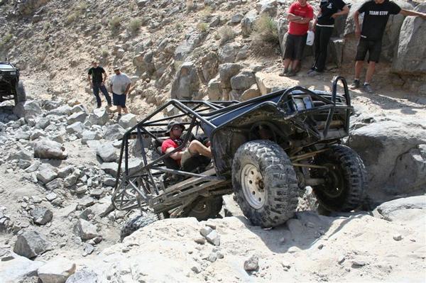 Black off-road buggy with roll cage navigating rocky terrain, driver wearing red shirt