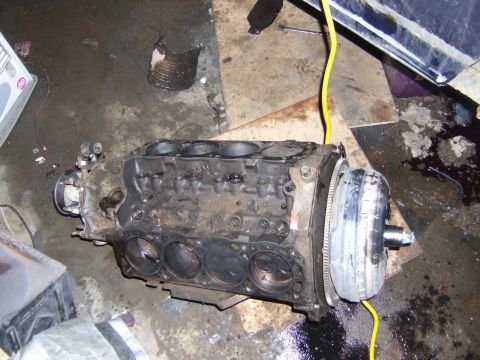 1989 Ford Bronco - An engine block and torque converter sit on a stained concrete floor, with a yellow cable running nearby. The Supermotors.net logo is visib…