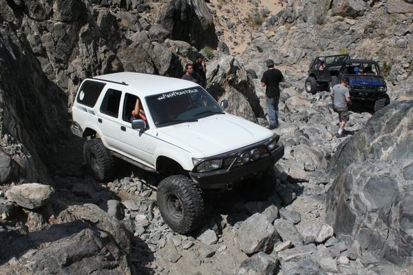 White 1993 Toyota 4Runner climbing over large rocks on an off-road trail. Two other off-road vehicles are visible further up the trail