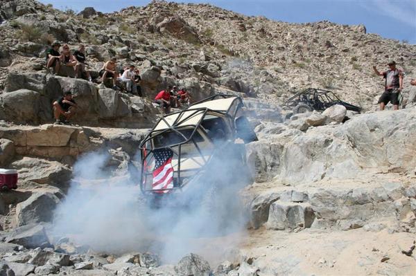 Off-road buggy with roll cage and American flag, tilted on rocks with smoke